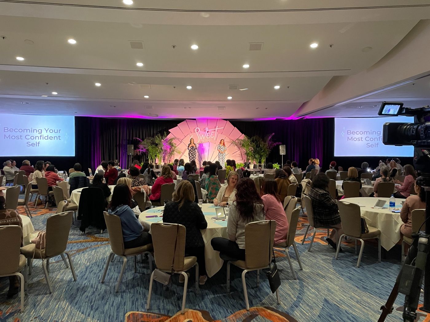 Women's retreat in a ballroom with a branded stage, two projection screens and a camera in the foreground